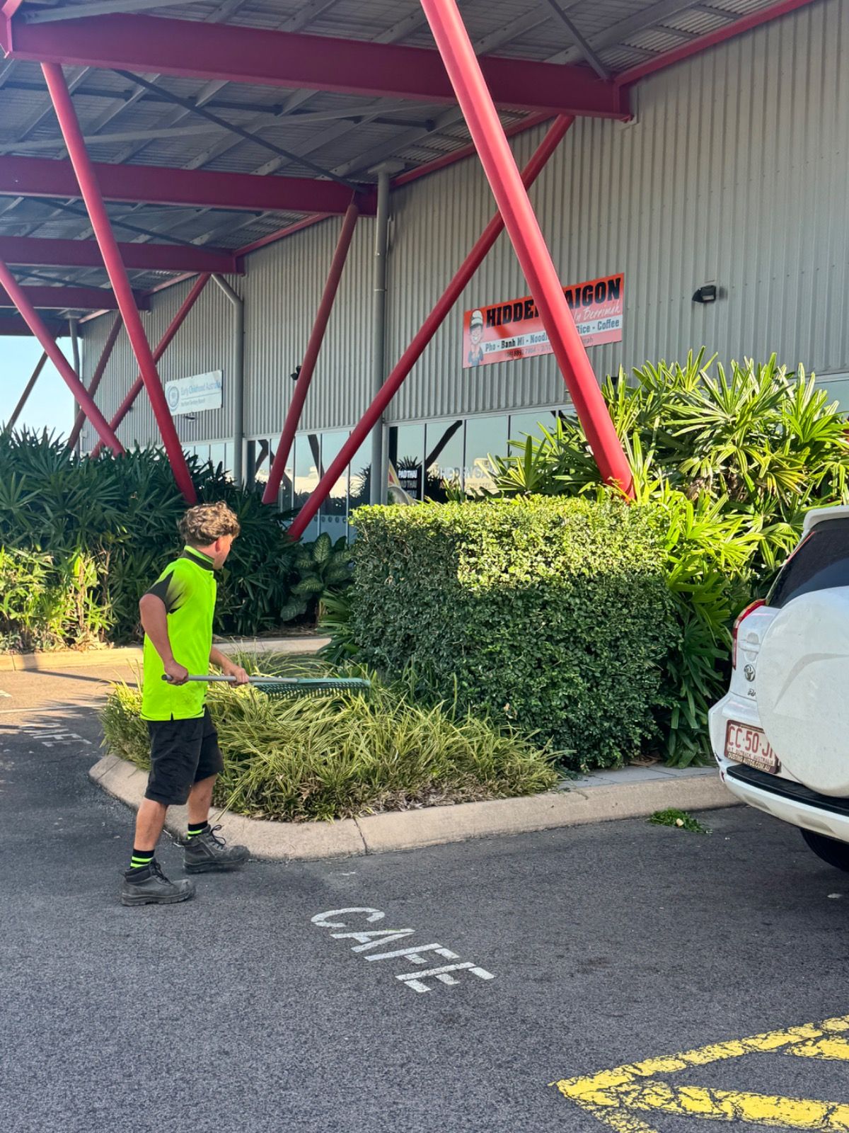 Gardener in bright yellow-green shirt trimming hedges near commercial building with red steel beams and surrounding shrubs in Berrimah NT